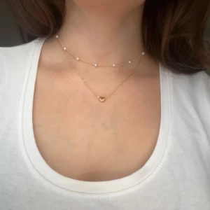 Dubbel halsband med hjärta och pärlor - Snyggt dubbel halsband med ett litet hjärta och små vita pärlor. Perfekt för att lägga till en subtil men elegant touch till din outfit. Passar både till vardags och festliga tillfällen. Halsbandet är i nytt skick och ger en fin lager-på-lager-effekt.