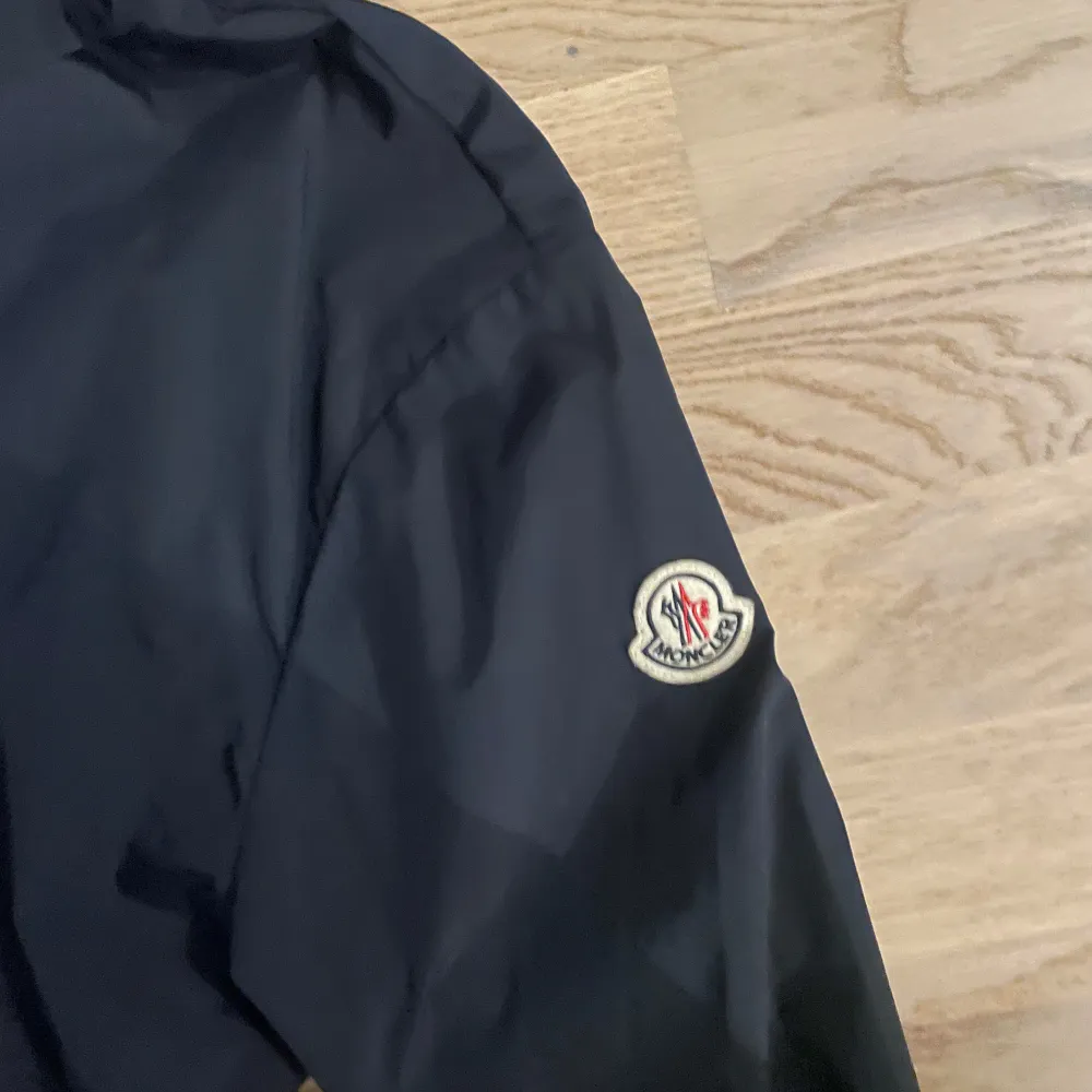 Säljer en snygg svart vindjacka från Moncler. Den har en dragkedja framtill och en huva med röd och vit randig detalj. Perfekt för höst och vår! Jackan har långa ärmar och en broderad Moncler-logga på ärmen. Passar både till vardags och lite finare tillfällen.. Jackor.