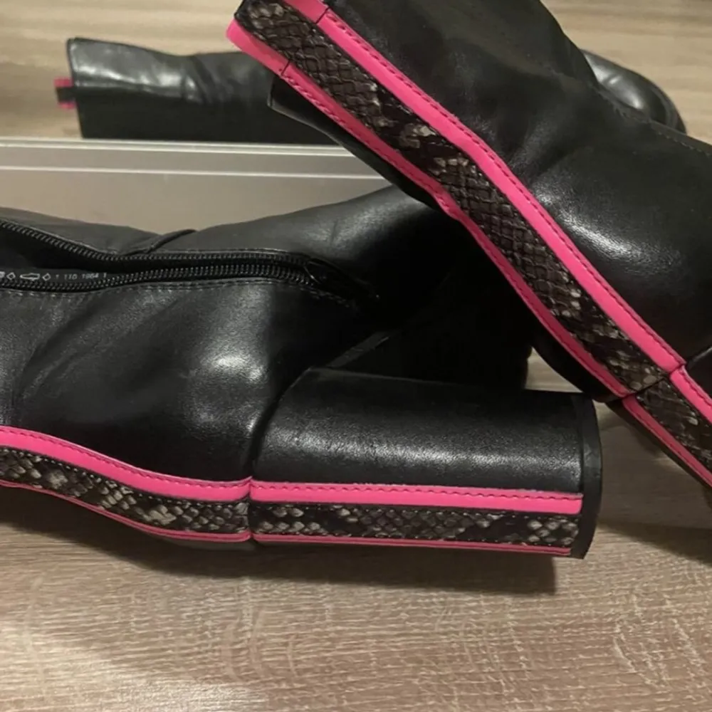 Snygga svarta boots med hög klack och coola rosa detaljer. De har en unik ormskinnsmönstrad rem längs baksidan och sulan, vilket ger dem en edgy look. Perfekta för höst och vinter, och de har en dragkedja på sidan för enkel på- och avtagning. Passar både till vardags och fest!. Skor.