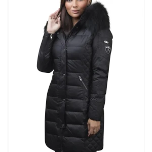 Svart lång dunjacka med pälsdetaljer - Säljer en elegant svart lång dunjacka med fluffig päls på huvan. Jackan har en quiltad design och dragkedjor på fickorna. Perfekt för kalla vinterdagar och håller dig varm och stilren. Äkta päls 