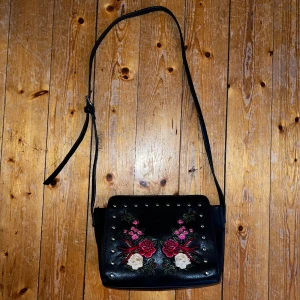 Svart axelväska med blommigt broderi - Säljer en superfin svart axelväska i skinnmaterial med vackert broderade blommor i rosa, rött och vitt. Väskan har en justerbar axelrem och är dekorerad med små nitar. Invändigt finns en praktisk dragkedjeficka för extra säkerhet. Köparen står för frakt, skickar spårtbart PostNord paket.