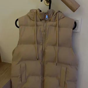 Beige dunväst med huva - Säljer en snygg beige dunväst med dragkedja och huva. Perfekt för kyliga dagar när du vill hålla värmen utan att bära en jacka. Västen har en stilren design med två fickor framtill. Passar bra till både vardag och utflykter.