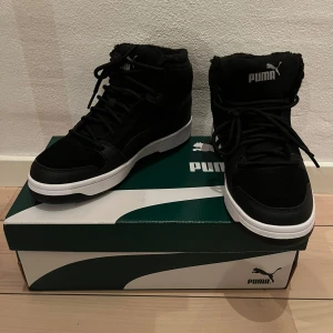 Svarta sneakers från Puma storlek 37. Nya  - Höga sneakers från Puma. Sneakers Rebound Layup Fur SD JR från Puma i mocka med detaljer i syntet och varmfodrade med mjuk fuskpäls. Plös och skaft är bekvämt vadderade.  Funktion: Varmfodrat Innersula: Pälsimitation Ovandel: Skinn/syntet Yttersula: Gummi