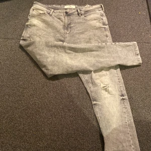 Grå jeans från FSBN - Säljer ett par grå jeans från FSBN i bra skick. De har en snygg, tvättad look med en liten reva på ena benet för en cool touch. Perfekta för vardagsbruk eller en avslappnad utekväll. Midjan är normal och passformen är slim, vilket ger en modern och stilren siluett.
