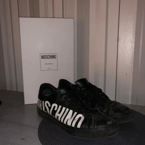 Svarta sneakers från Moschino - Säljer ett par svarta Moschino sneakers i skinn med stort vitt Moschino-tryck på sidan. Skorna har snörning och är i bra skick. Perfekta för en stilren look med en touch av lyx. Kommer med originalkartong. Nypris runt 3000kr🙌