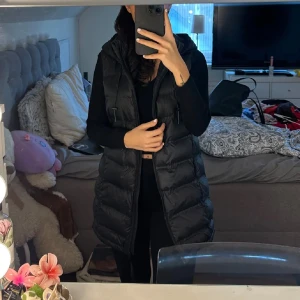 Svart lång dunväst - Säljer en svart lång dunväst i mycket bra skick. Västen har en dragkedja och är perfekt för höst och vinter. Den är stilren och passar till många olika outfits. Perfekt för att hålla sig varm och snygg under kyliga dagar. Använd två tre gånger passa även S 