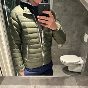 Grön dunjacka från Sail Racing - Säljer en grön dunjacka från Sail Racing i mycket bra skick. Jackan har en snygg pufferstil med dragkedja och är perfekt för höst och vinter. Den är långärmad och har en modern look som passar både till vardags och lite finare tillfällen.