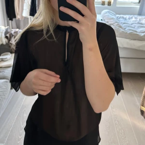 Svart blus med knapp - Säljer en elegant svart blus i mycket bra skick. Den har korta ärmar och en knapp vid halsen. Perfekt för både vardag och festliga tillfällen. Materialet är lätt och luftigt, vilket gör den bekväm att bära hela dagen. Märket är Gina Tricot och storlek 36