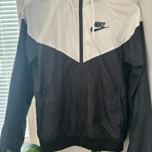 Svart och vit vindjacka från Nike - Säljer en snygg svart och vit vindjacka från Nike i storlek XS men skulle säga att den också passar för storlek S och kanske M. Jackan har en dragkedja framtill och en huva för extra skydd mot väder. Perfekt för vår och höst! Den är i mycket bra skick och redo för nya äventyr och funkar för båda killar och tjejer. Den är knappt använd. 