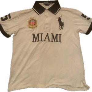 Miami Ralph Lauren Polo - Säljer en snygg vit pikétröja från Ralph Lauren med 'MIAMI' tryckt på framsidan och en stor siffra 5 på baksidan. Tröjan har korta ärmar och en klassisk krage med knappar. Perfekt för en avslappnad men stilren look!