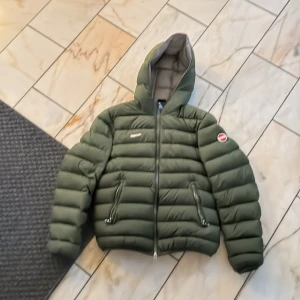 Grön dunjacka från Colmar - Säljer en snygg grön dunjacka från Colmar i mycket bra skick. Jackan har en pufferstil med dragkedja och huva, perfekt för kyliga dagar. Den är långärmad och har en normal passform. Märkeslogga på ärmen ger en extra touch. Perfekt för höst och vinter! Säljer billigt pågrund av en tejpbit som sitter fast på jackan. Strl 152 men passar upp till 160