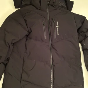 Svart pufferjacka från Sail Racing - Säljer en svart pufferjacka från Sail Racing i bra skick. Jackan har en dragkedja framtill och två bröstfickor med dragkedjor. Perfekt för kalla vinterdagar. Det finns en liten skada på båda ärmarna, men inget som påverkar funktionen. Passar både till vardags och utomhusaktiviteter. Storleken är 150cm men modellen på bilden är 170cm! Ny pris 3500 mitt pris 1200🤩
