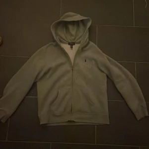 Grå hoodie från Polo Ralph Lauren - Säljer en stilren grå hoodie från Polo Ralph Lauren. Den är barn Xl men passar som M/S i vuxen storlek. Den har en dragkedja framtill och en klassisk logga på bröstet. Perfekt för både höst och vår, och den är supermjuk och bekväm. Passar till alla tillfällen!