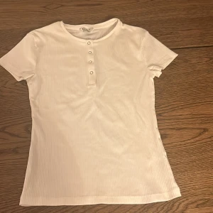 Vit ribbad topp i ekologisk bomull - Säljer en vit ribbad topp i ekologisk bomull från märket Cubus. Toppen har korta ärmar och knappar framtill, perfekt för en avslappnad stil. Den är i mycket bra skick och passar perfekt till både vardag och fest.