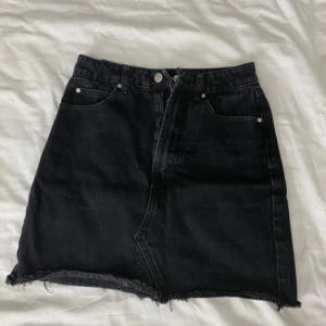 Svart jeanskjol - Säljer en svart jeanskjol i kort modell med rå kant och klassisk femficksdesign. Perfekt för en avslappnad look, passar både till vardags och fest. Dragkedja och knapp framtill för enkel påklädning. Skickar gärna fler bilder vid intresse!