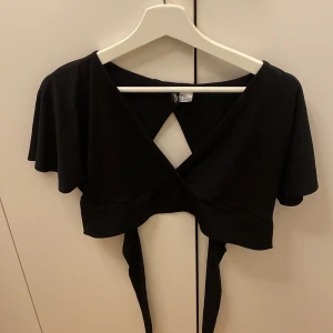 Svart omlottopp från H&M Divided - Snygg svart omlottopp från H&M Divided i storlek M. Toppen har korta ärmar och en knytning i ryggen som ger en fin detalj. Perfekt för både vardag och fest!