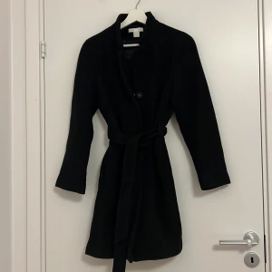 Svart kappa i ullblandning - Säljer en elegant svart kappa från H&M i ullblandning. Den har en klassisk design med knappar och ett bälte i midjan för en snygg passform. Perfekt för höst och vinter, med långa ärmar som håller dig varm. Passar både till vardags och festliga tillfällen.