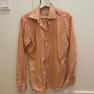 Orange och vit randig skjorta från Eton - Säljer en snygg orange och vit randig skjorta från Eton i storlek 40. Skjortan har en slim passform och är perfekt för både vardag och fest. Den är långärmad och har klassiska knappar framtill. Materialet känns skönt och andas bra. Perfekt för vår och sommar! 🌞