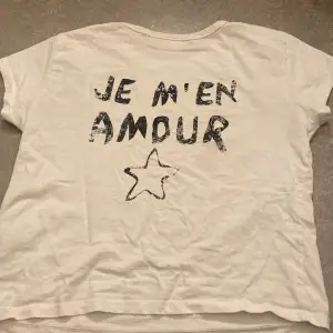 Säljer en vit t-shirt från Gina Tricot med texten 'JE M'EN AMOUR' och en stjärna tryckt på framsidan. T-shirten är kortärmad, i bomull och i storleken 158/164. Tvättning och strykning sker innan packning till köparen 💘💕💗🩷