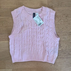 Rosa stickad väst  - Säljer en superfin rosa stickad väst. Den har ett klassiskt flätmönster och ribbade kanter. Perfekt för lager-på-lager under hösten eller våren. Helt ny med lapp kvar 