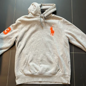 Grå hoodie från Ralph Lauren - Säljer en snygg grå hoodie från Ralph Lauren med orange logga på bröstet och siffran 3 på ärmen. Perfekt för en casual look och superbekväm att ha på sig. Den har en klassisk känguruficka och justerbar huva. Passar perfekt till jeans eller joggers! Storlek är L passar M-L. Skick 9/10