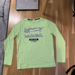 Limegrön långärmad tröja från Lindex - Säljer en cool limegrön långärmad tröja från Lindex med ett tryck av New York-silhuetten och texten 'New York My World'. Perfekt för vardagsbruk och att sticka ut med sin unika färg. Passar ungdomar i åldern 12-14 år.