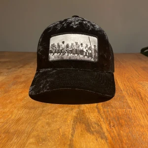 Svart keps från Hatstore - Snygg svart keps från Hatstore med ett coolt tryck av ett klassiskt foto på framsidan. Perfekt för att ge din outfit en unik touch. Kepsen har en justerbar passform och är i mycket bra skick. Jag har inte använt den så mycket.