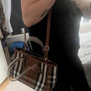 Säljer en snygg rutig handväska med bruna läderdetaljer och dragkedja. Väskan har ett klassiskt rutmönster i beige, svart och rött. Perfekt för både vardag och fest! 👜✨