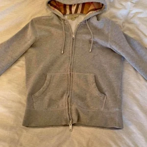 Grå hoodie från Burberry - Säljer en snygg grå hoodie från Burberry med klassiskt rutigt mönster i luvan. Den har dragkedja framtill och två praktiska fickor. Perfekt för höst och vår när du vill vara både stilren och bekväm. Passar till alla tillfällen! Säljer min burberry zip eftersom den är för liten för mig. Den är storlek S. Den har inga defekter. Om du är intresserad är det bara att höra av dig. 