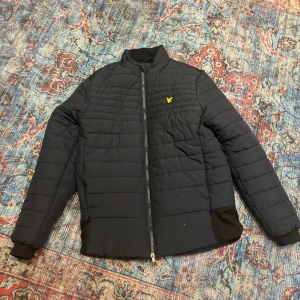 Svart pufferjacka från Lyle & Scott - Säljer en mörkblå pufferjacka från Lyle & Scott i mycket bra skick. Jackan har en dragkedja framtill och är långärmad med ribbade muddar. Perfekt för höst och vinter, med en stilren design och det klassiska Lyle & Scott-emblemet på bröstet. Superbekväm och varm!