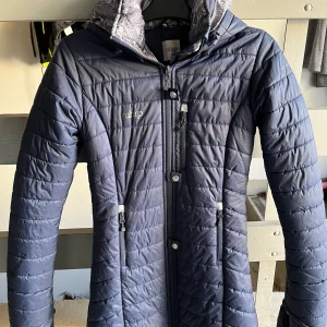 Mörkblå quiltad jacka från Uhip - Säljer en snygg mörkblå quiltad jacka från Uhip i storlek 150. Jackan har en dragkedja och knappar framtill, samt en mysig huva med mönstrat foder. Perfekt för kyliga höst- och vinterdagar. Den är i mycket bra skick nästan aldrig använd och redo för nya äventyr!