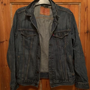 Jeansjacka från Levi's - Säljer en klassisk jeansjacka från Levi's i storlek M. Den är i bra skick och har en snygg mörkblå färg. Jackan har långa ärmar och två bröstfickor med knappar. Perfekt för vår och höst!
