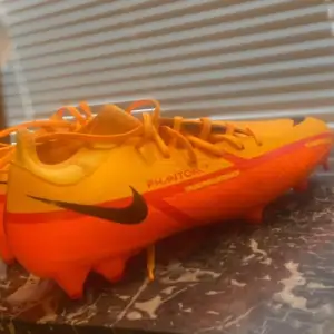 Säljer ett par Nike fotbollsskor i en cool orange och gul färgkombination. Skorna har snörning och är i nytt skick. Perfekta för fotbollsträningen med en snygg design och bra grepp på planen.