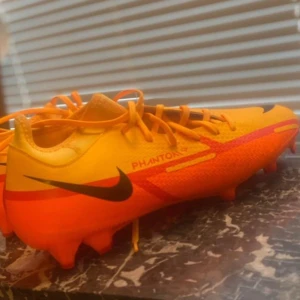 Nike fotbollsskor i orange och gul - Säljer ett par Nike fotbollsskor i en cool orange och gul färgkombination. Skorna har snörning och är i nytt skick. Perfekta för fotbollsträningen med en snygg design och bra grepp på planen.