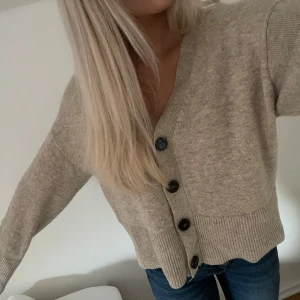 Beige kofta från Stockh LM - Mysig beige kofta från Stockh LM i storlek S. Den har en v-ringad design med stora knappar framtill och ribbade muddar. Perfekt för höst och vår när du vill ha något bekvämt och stilrent. Passar både till jeans och kjol!