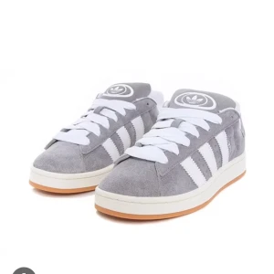 Grå Adidas sneakers - Snygga grå Adidas sneakers i mocka med vita detaljer och klassiska ränder. De har en bekväm passform och vit sula med gummidetaljer. Perfekta för vardagsbruk eller en avslappnad stil. Skorna har snörning för en justerbar passform.