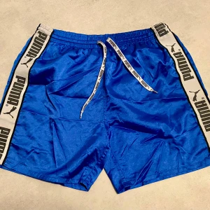 Blå shorts från Puma - Säljer ett par blå Puma shorts i mycket bra skick. De har en elastisk midja med snörning och sidopaneler med Puma-logga. Perfekta för träning eller en avslappnad dag. Några mindre fläckar finns, men inget som sticker ut. Skönt glansigt material! Storlek S men passar även M bra.