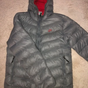 Grå dunjacka med röd insida storlek M/L - Säljer en snygg grå dunjacka från Nike med en cool röd insida. Jackan har en dragkedja framtill och två praktiska fickor med dragkedjor. Perfekt för kalla dagar med sin varma och puffiga design. Passar både till vardags och utomhusaktiviteter.