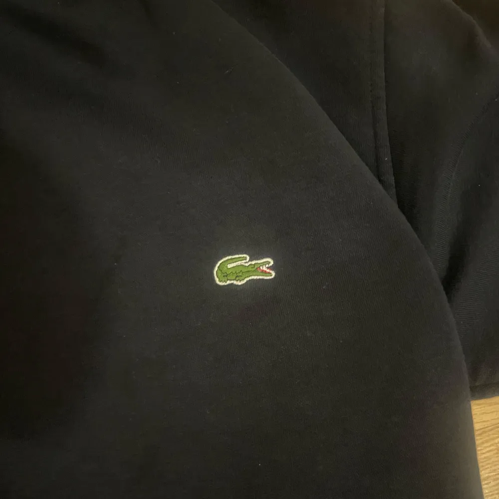 Säljer en snygg mörk hoodie från Lacoste med den klassiska krokodilloggan på bröstet och Lacoste-text på ärmen. Perfekt för en casual look eller att mysa i hemma. Den har en stor ficka fram och en bekväm huva. Passar till alla tillfällen!. Hoodies.