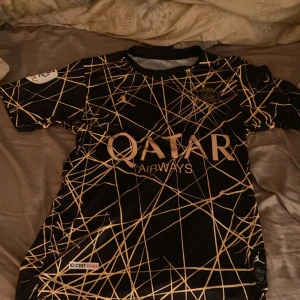 Svart och gul PSG fotbollströja - Säljer en snygg PSG fotbollströja i svart med guldmönster. Tröjan har korta ärmar och är i mycket bra skick. Perfekt för alla PSG-fans som vill sticka ut på planen eller på stan. Den har detaljer som 'Qatar Airways' och 'Ici C'est Paris'.
