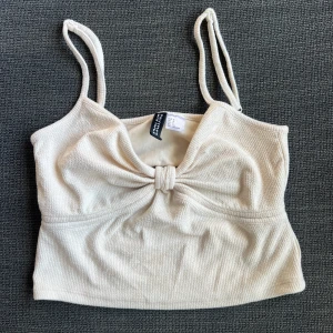 Beige ribbad topp  - Säljer en superfin beige ribbad topp från Divided. Den har smala axelband och en söt rosett framtill. Toppen är croppad och perfekt för sommardagar. Baktill finns en liten öppning som ger en extra touch. Perfekt för en avslappnad och stilren look!