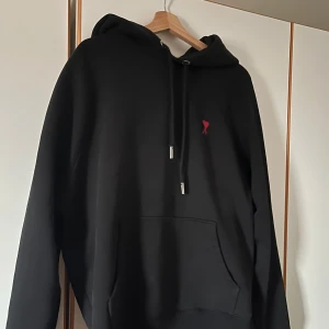 Svart hoodie från Ami - Säljer en stilren svart hoodie från Ami med en liten röd logga på bröstet. Den har justerbar huva med snören.