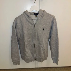 Grå hoodie från Polo Ralph Lauren - Säljer en snygg och bekväm grå hoodie från Polo Ralph Lauren. Den har en dragkedja framtill och två praktiska fickor. Perfekt för en avslappnad stil och passar till de flesta tillfällen. 🐎