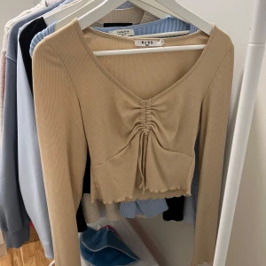 Beige ribbad topp från NA-KD - Snygg beige ribbad topp från NA-KD med v-ringning och knytning framtill. Den har långa ärmar och är croppad, perfekt för både vardag och fest. Toppen är i mycket bra skick och passar perfekt till höstgarderoben.
