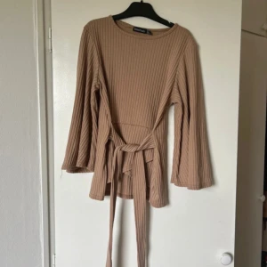 Beige ribbad tröja från Boohoo - Säljer en superfin beige ribbad tröja från Boohoo. Den har långa ärmar och ett knytband i midjan som ger en snygg passform. Perfekt för höst och vår! Tröjan är i mycket bra skick och passar både till vardags och lite finare tillfällen.