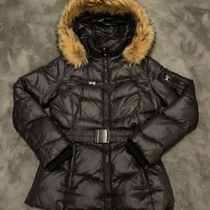 Svart pufferjacka med pälsdetaljer - Säljer en snygg svart pufferjacka med fuskpäls på huvan. Jackan har ett bälte i midjan och dragkedja framtill. Perfekt för kalla vinterdagar! Den har långa ärmar och flera praktiska fickor. Håll dig varm och stilren i vinter! 🖤