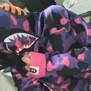 Säljer en cool hoodie från BAPE med ett unikt hajmotiv på luvan. Den har ett färgglatt camouflagemönster i lila, rosa och svart. Hoodien är långärmad med ribbade muddar och dragkedja. Perfekt för att sticka ut i mängden! Mycket bra skick priset kan diskuteras 