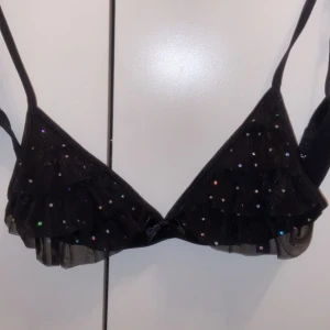 Svart glittrig bh med en rosett från Lindex - Snygg svart bh från Lindex med glittriga paljetter och volangdetaljer. Storlek M (40/42). Mycket bra skick!