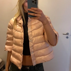 Ljusrosa pufferjacka - Säljer en superfin ljusrosa pufferjacka med trekvartsärm. Den har stora knappar framtill och en glansig finish som ger en lyxig känsla. Perfekt för vår och höst när det är lite kyligt ute. Jackan är i mycket bra skick.