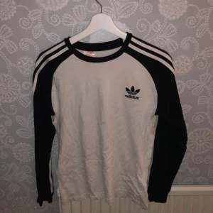 Vit och svart långärmad tröja från Adidas - Säljer en snygg vit och svart långärmad tröja från Adidas. Den har de klassiska tre ränderna längs ärmarna och Adidas-loggan på bröstet. Perfekt för en sportig look eller vardagsstil. Passar bra till både jeans och träningsbyxor. Hör av dig vid intresse!